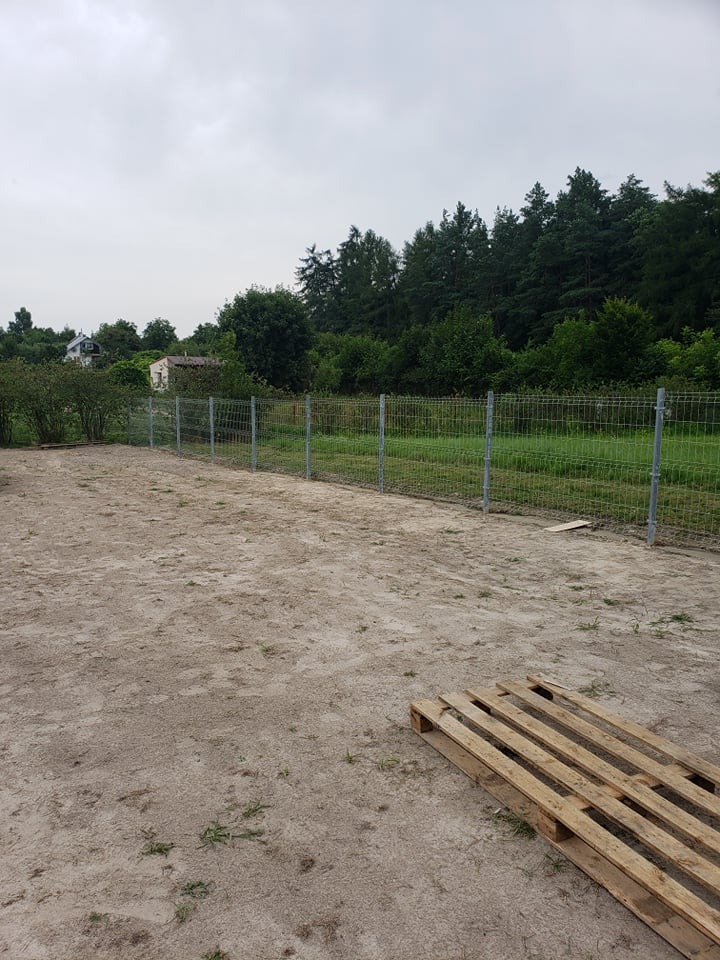 Nowo postawione ogrodzenie z siatki na metalowych słupkach, widoczne na piaszczystym terenie z paletą w rogu, w tle zieleń drzew i krzewów pod pochmurnym niebem.