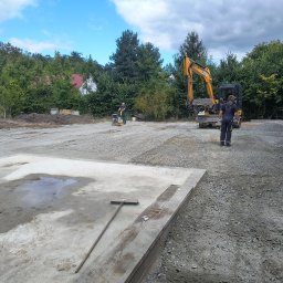 Mi-kop - Budowa fundamentów: betonowa płyta z kałużą, ubijarka i koparka JCB w tle, przygotowany teren pod budowę, częściowo wysypany gruzem, rusztowania przy budynku.