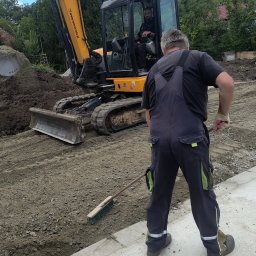 Mi-kop - Żółta koparka JCB i pracownik z miotłą na placu budowy. Operator w kabinie koparki. Przygotowanie terenu pod budowę, wyrównywanie ziemi.