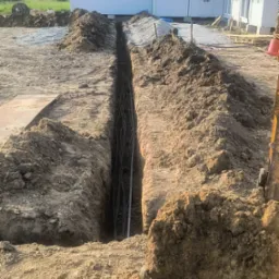 Długi wykop w ziemi z ułożonymi kablami, przygotowany do zasypania. W tle kontenery biurowe i hałda żwiru. Widoczne ślady pracy maszyny budowlanej.