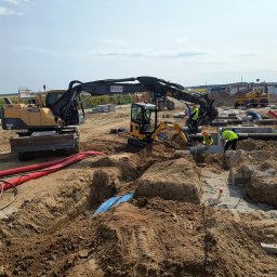 Mi-kop - Prace ziemne z użyciem koparek Volvo i JCB. Widoczne wykopy, fundamenty, rury instalacyjne. Pracownicy w żółtych kamizelkach. Słoneczny dzień, plac budowy.