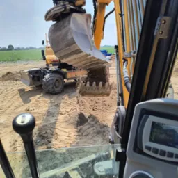 Żółta koparka w akcji, z perspektywy operatora, wysypuje ziemię na placu budowy. W tle widoczna zielona łąka i błękitne rury.