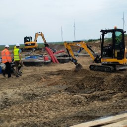 Mi-kop - Żółty minikoparka JCB na placu budowy z widocznymi rurami i kablami. W tle turbiny wiatrowe i dwóch robotników w kaskach. Prace ziemne w toku.