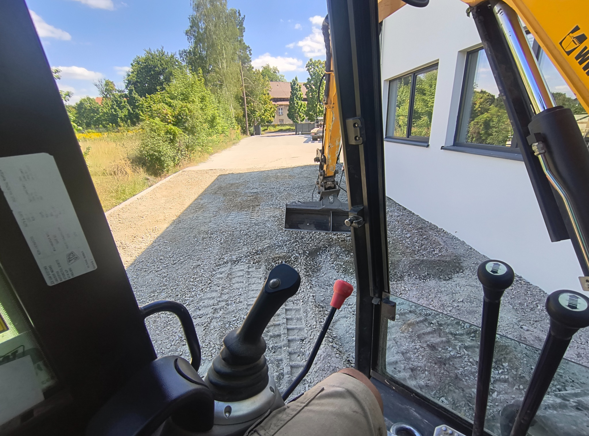 Widok z kabiny koparki na utwardzony żwirem teren, zarys budynku w tle. Operator w kabinie, widoczne elementy sterowania. Słoneczny dzień, perspektywa z wnętrza maszyny.