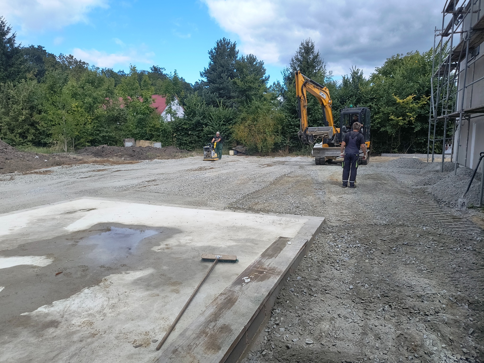 Budowa fundamentów: betonowa płyta z kałużą, ubijarka i koparka JCB w tle, przygotowany teren pod budowę, częściowo wysypany gruzem, rusztowania przy budynku.