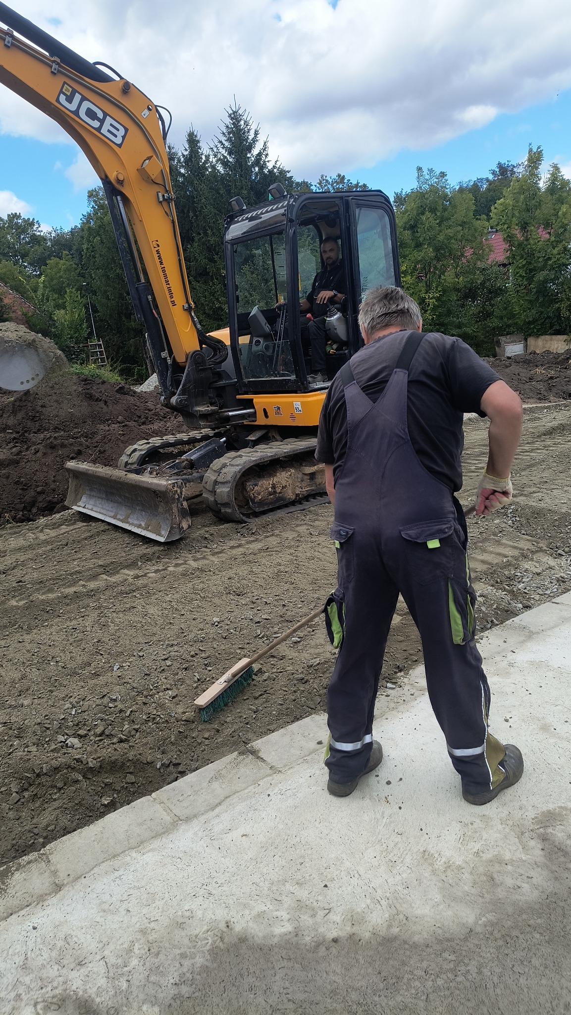 Żółta koparka JCB i pracownik z miotłą na placu budowy. Operator w kabinie koparki. Przygotowanie terenu pod budowę, wyrównywanie ziemi.
