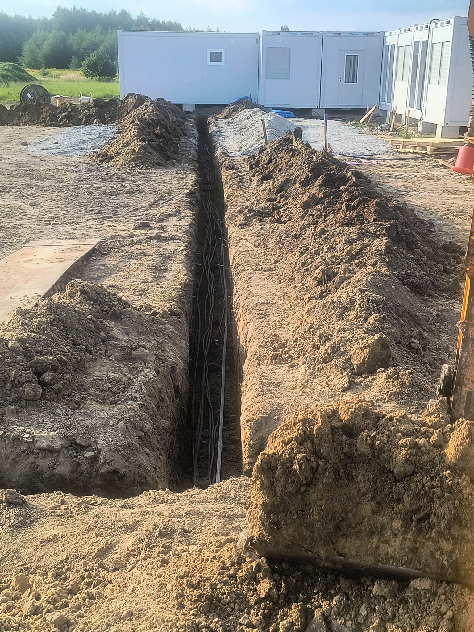 Długi wykop w ziemi z ułożonymi kablami, przygotowany do zasypania. W tle kontenery biurowe i hałda żwiru. Widoczne ślady pracy maszyny budowlanej.