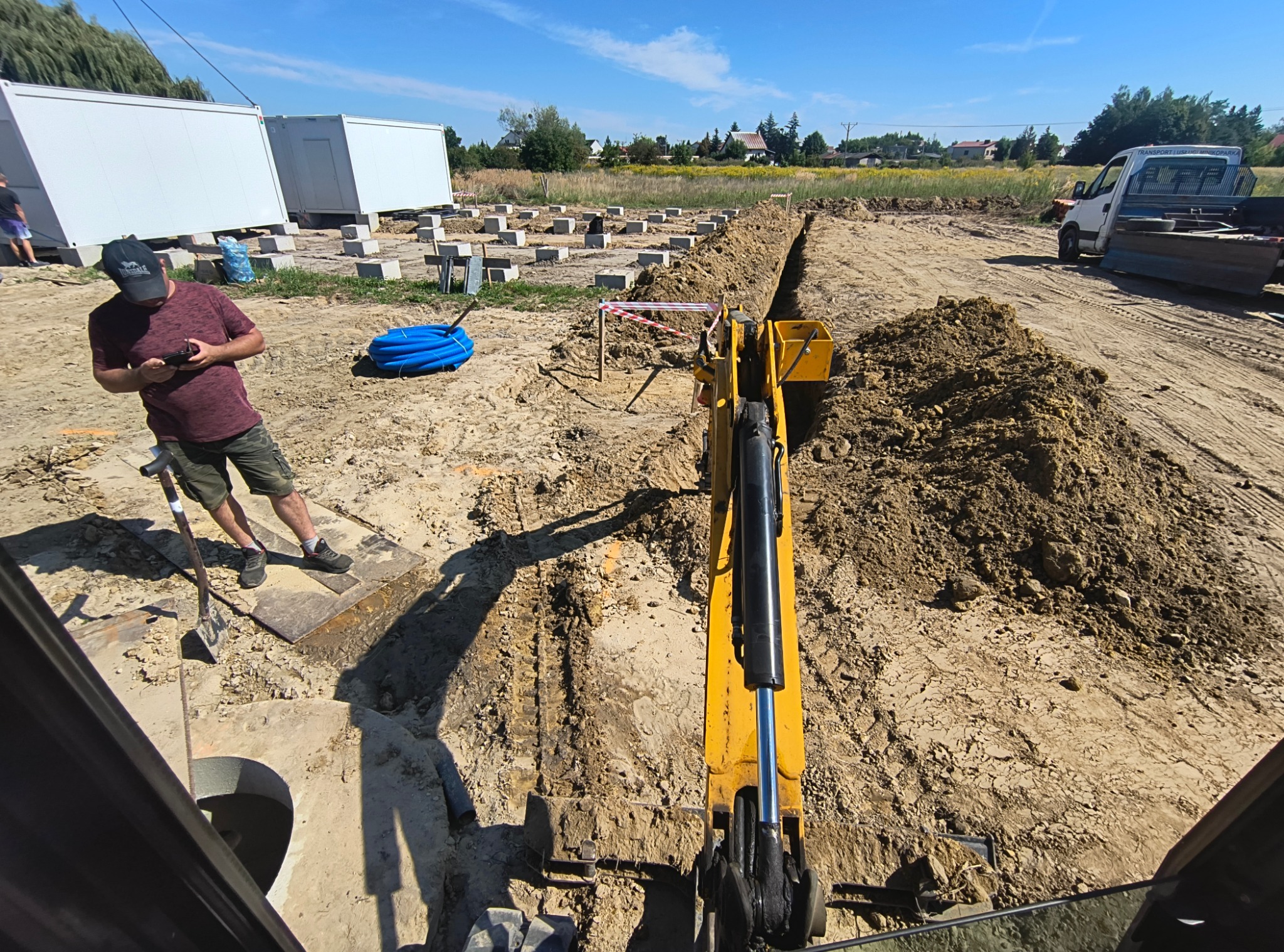 Żółta koparka w trakcie wykopu rowu na placu budowy z fundamentami. Operator z pilotem, widoczne kontenery i auto dostawcze w tle, słoneczny dzień.