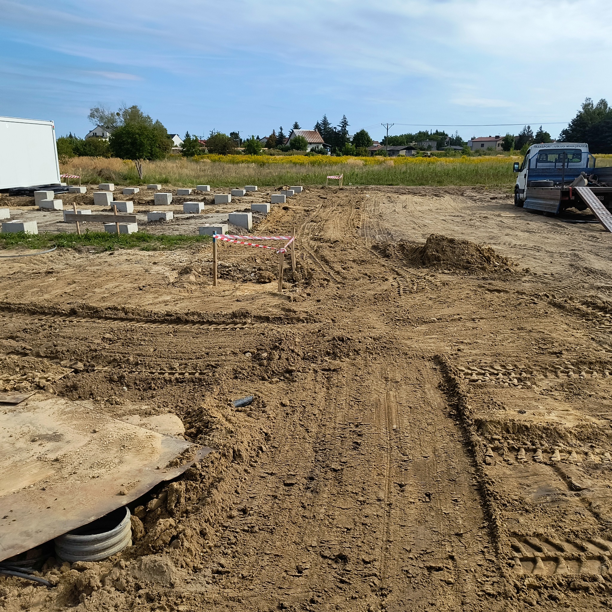 Przygotowany teren pod budowę z betonowymi bloczkami, wytyczone ścieżki i taśmy ostrzegawcze, biały kontener i samochód dostawczy w tle.
