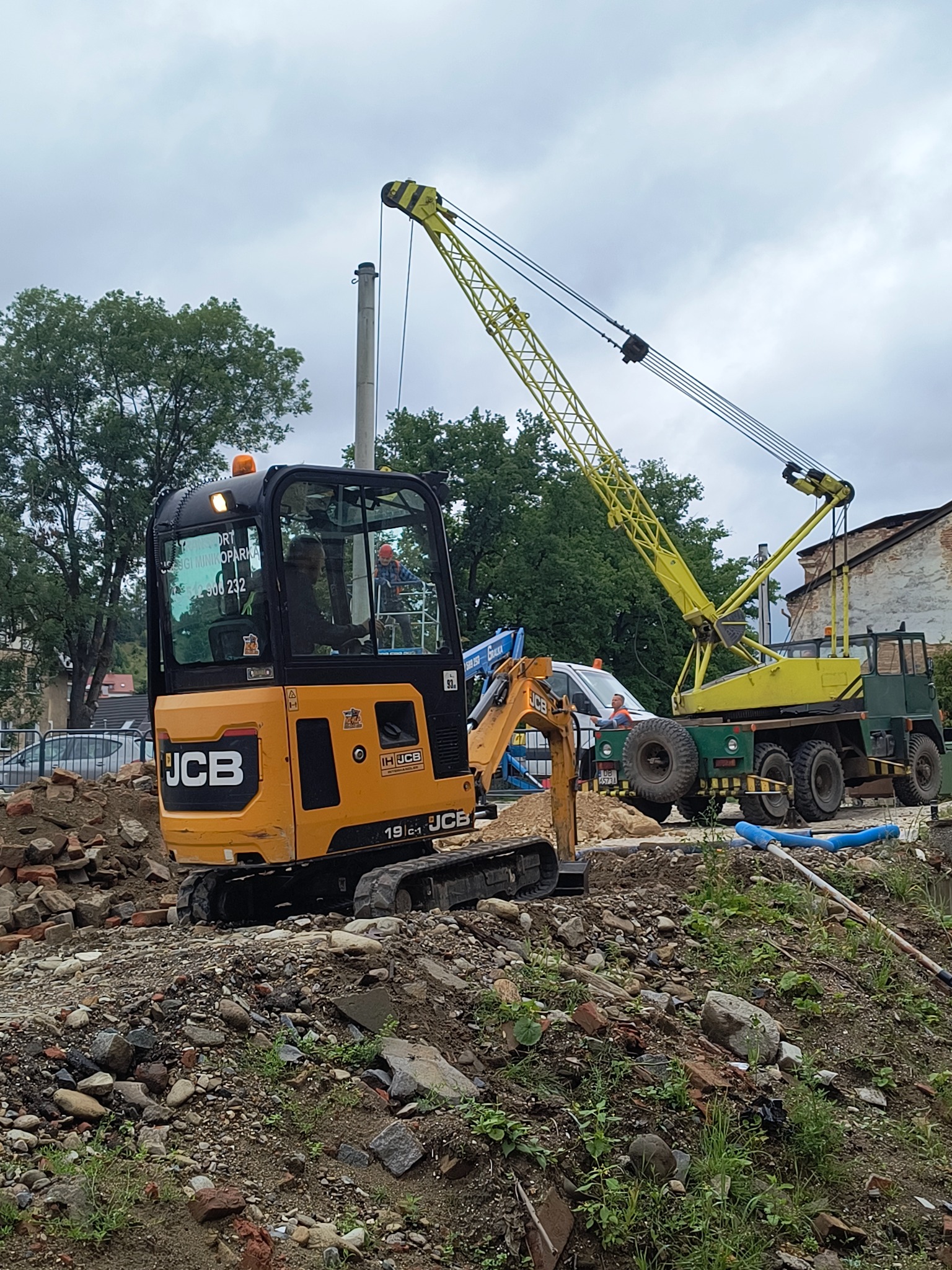 Żółta mini koparka JCB 19C-1 na placu budowy z gruzem i kamieniami, w tle dźwig samochodowy. Roboty ziemne w toku.