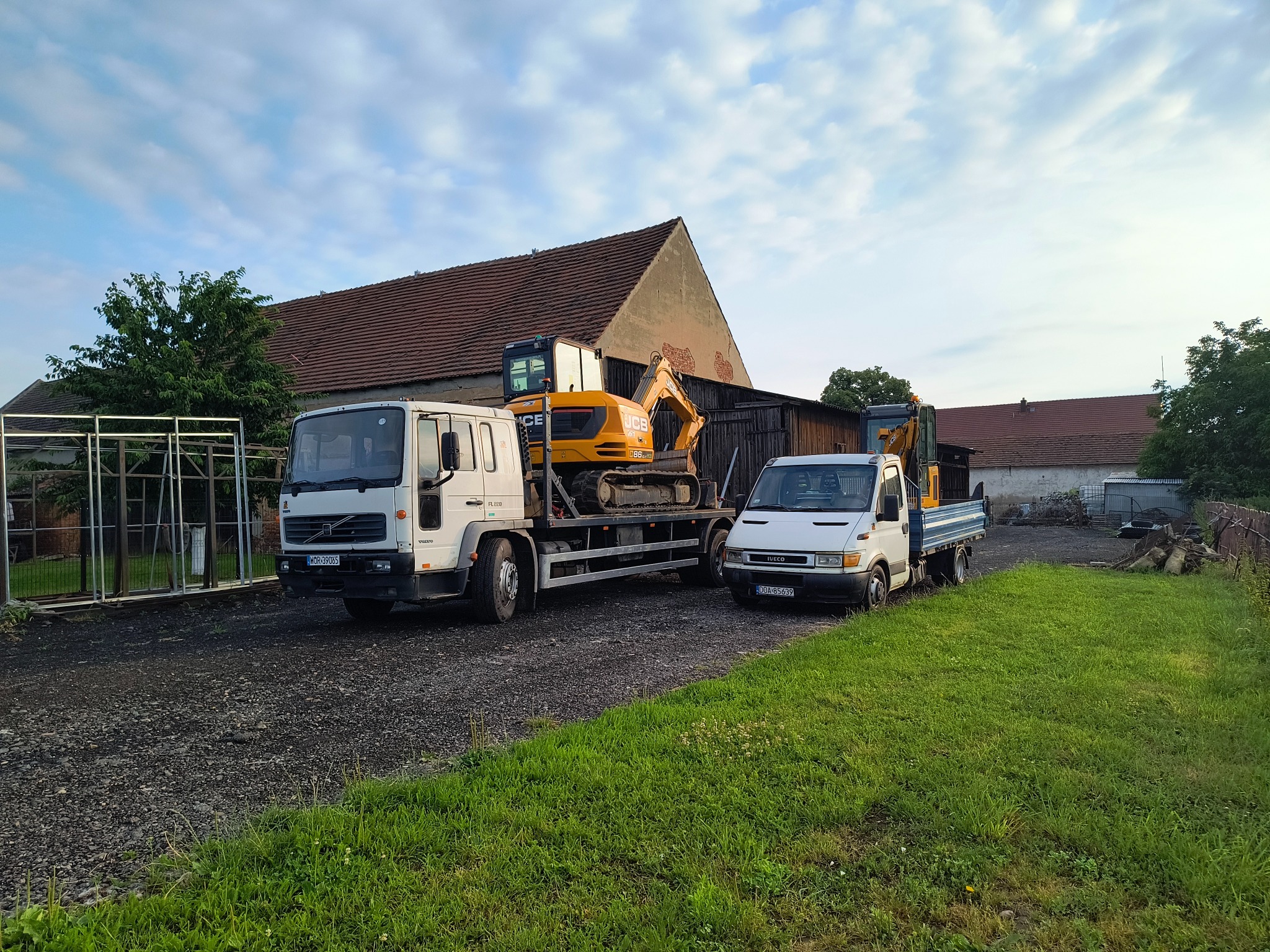 Dwa samochody ciężarowe, w tym jeden z żółtą koparką JCB na platformie, zaparkowane na żwirowym podłożu z trawą, na tle budynków gospodarczych.