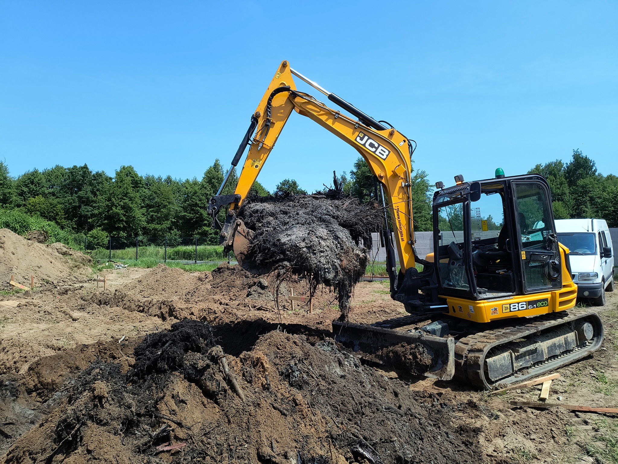 Żółta mini koparka JCB podnosi łyżką ziemię z korzeniami na tle wykopu i zieleni. W tle widoczny biały samochód dostawczy i błękitne niebo.