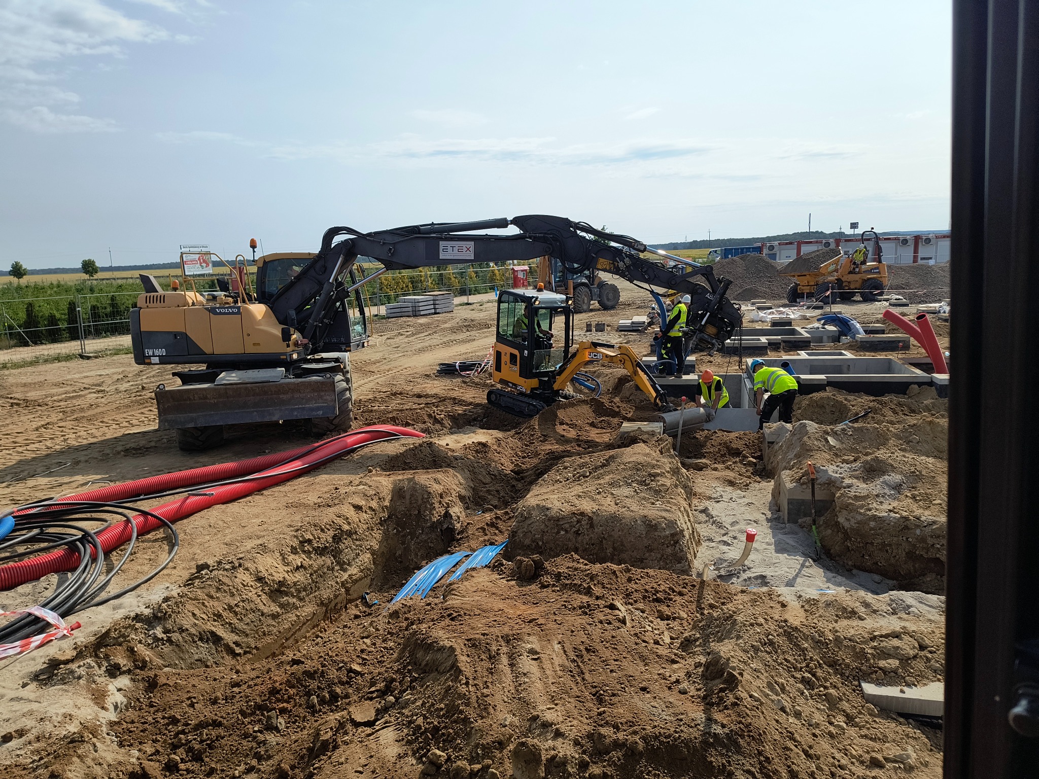Prace ziemne z użyciem koparek Volvo i JCB. Widoczne wykopy, fundamenty, rury instalacyjne. Pracownicy w żółtych kamizelkach. Słoneczny dzień, plac budowy.