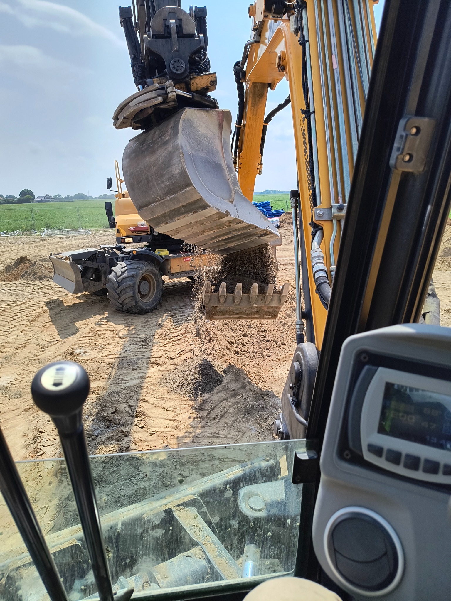 Żółta koparka w akcji, z perspektywy operatora, wysypuje ziemię na placu budowy. W tle widoczna zielona łąka i błękitne rury.