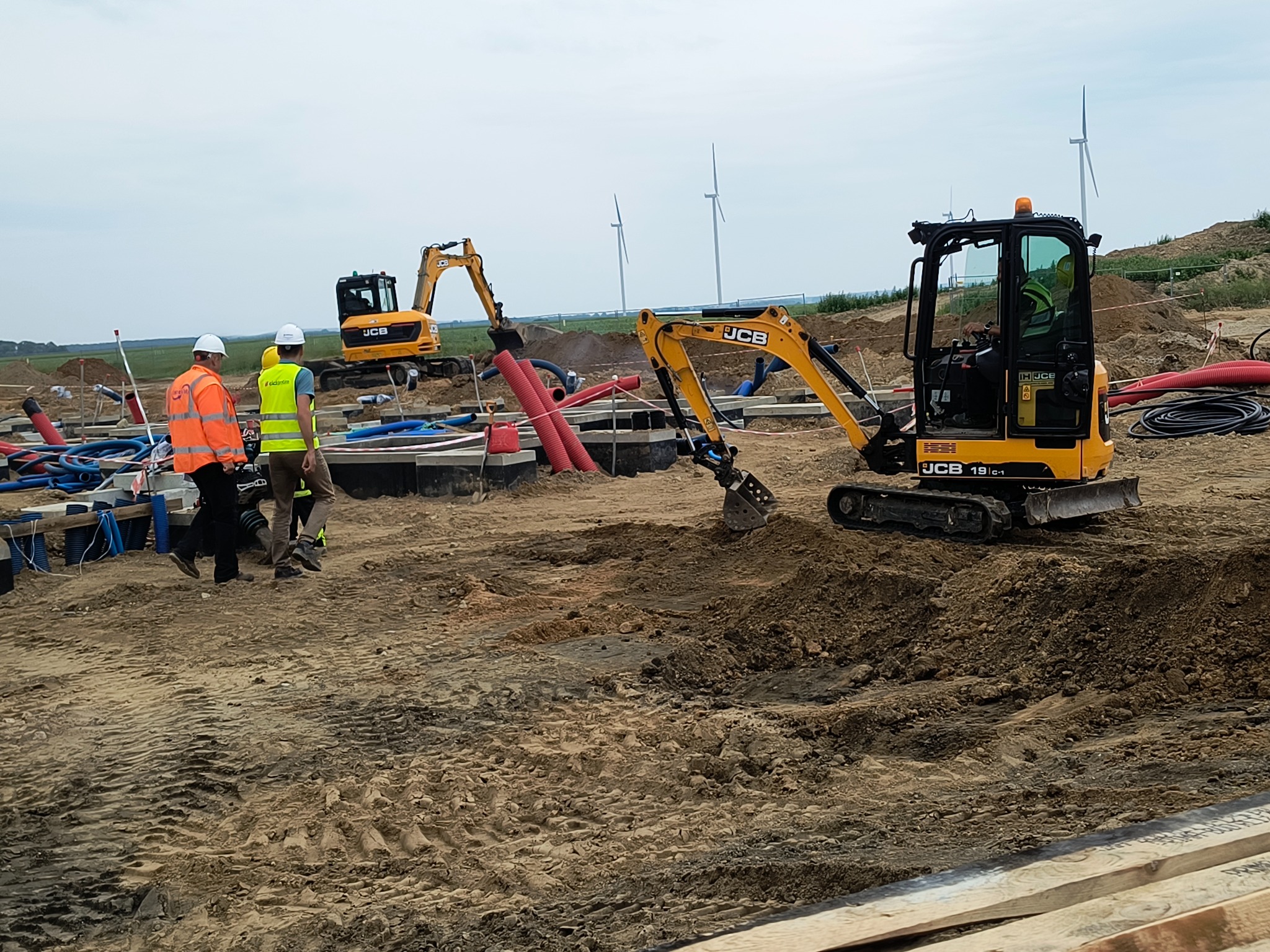 Żółty minikoparka JCB na placu budowy z widocznymi rurami i kablami. W tle turbiny wiatrowe i dwóch robotników w kaskach. Prace ziemne w toku.