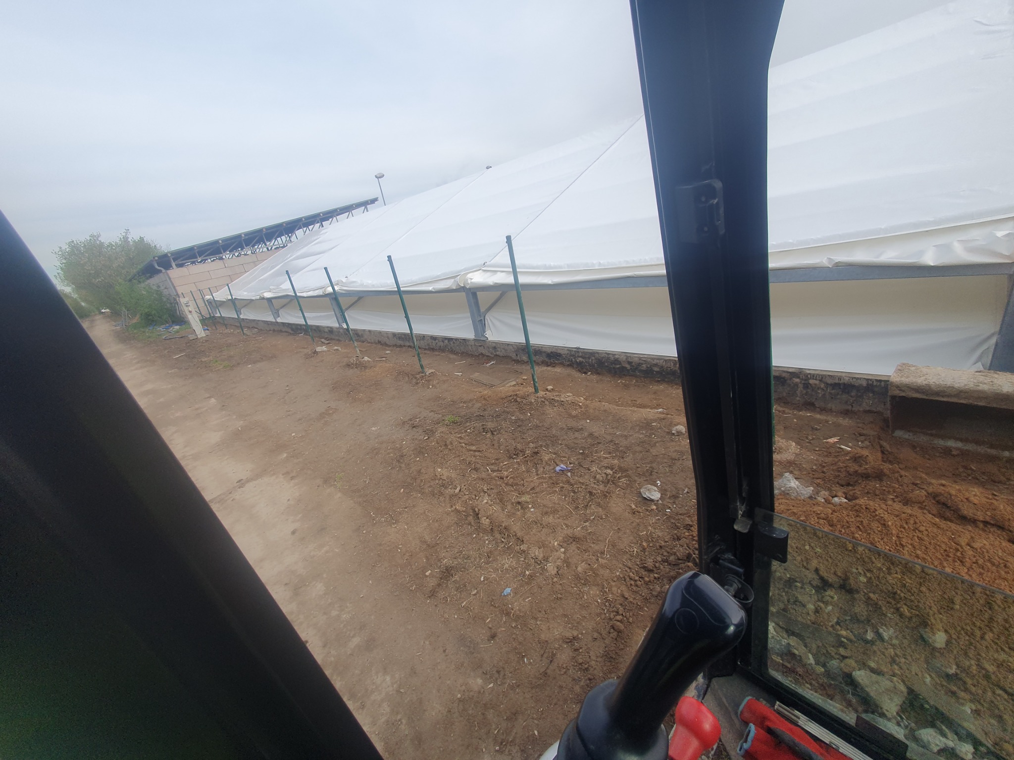 Widok z kabiny maszyny budowlanej na teren z przygotowanym podłożem i konstrukcją namiotową w tle, pochmurne niebo.