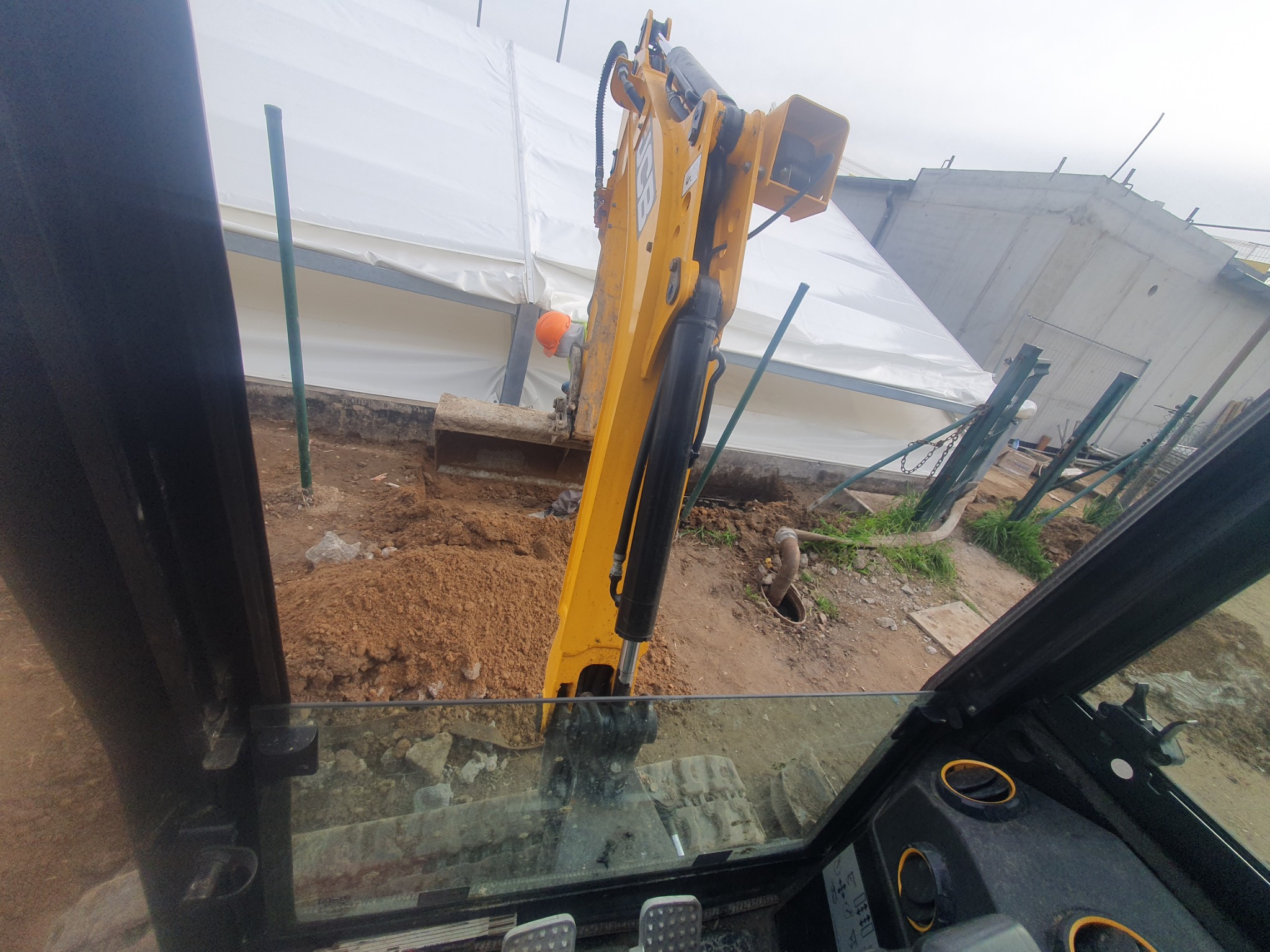 Widok z kabiny operatora żółtej koparki JCB podczas wykopu pod fundamenty, w tle biały namiot i betonowy budynek.