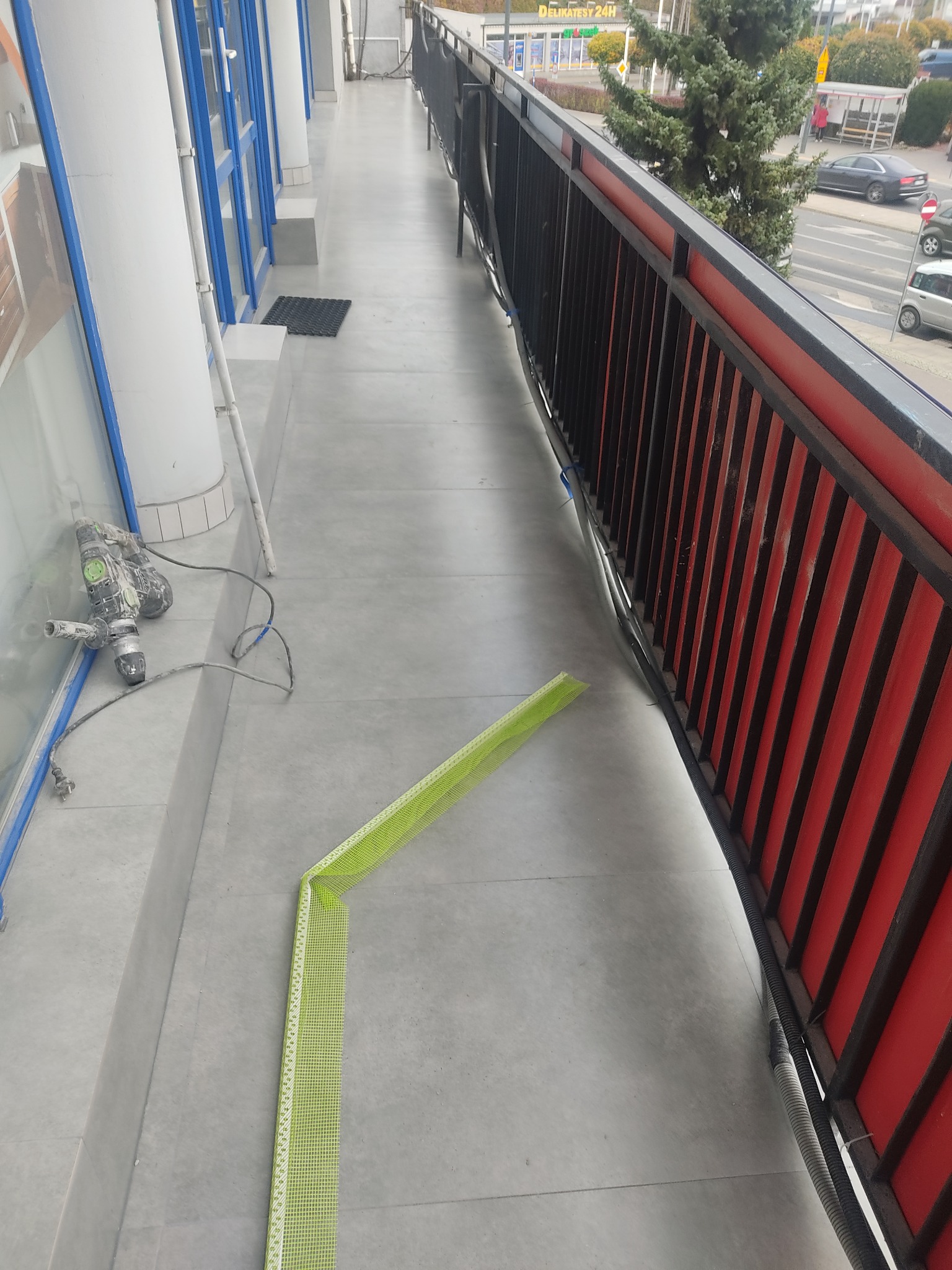 Szary balkon z nową, jasną, kwadratową płytką, zielona taśma pomiarowa wyznacza kąt, obok leży używana wiertarka udarowa, widok z góry na fragment ulicy.