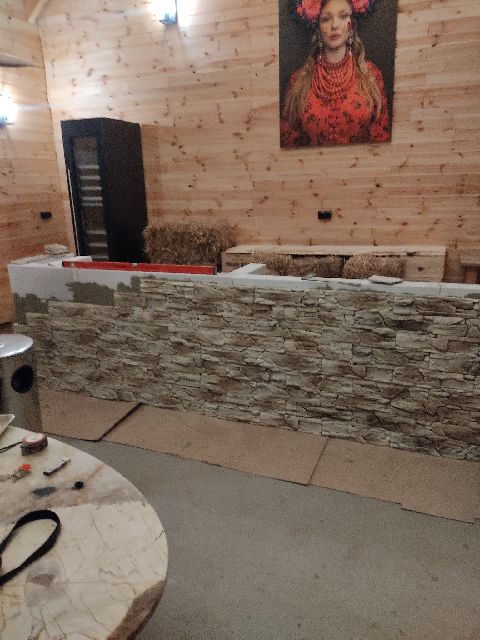 Montaż dekoracyjnej ściany z kamienia na zapleczu baru, widoczny poziomica, narzędzia na marmurowym stole i portret kobiety w ukraińskim stroju na ścianie z boazerii.