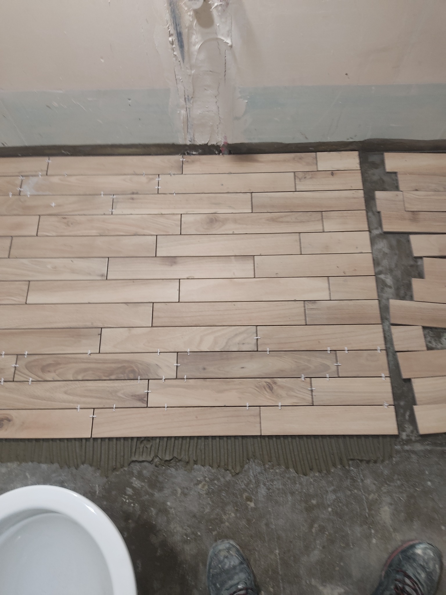 Układanie podłużnych płytek imitujących drewno w łazience, widoczne krzyżyki dystansowe, fragment toalety i buty osoby wykonującej prace.