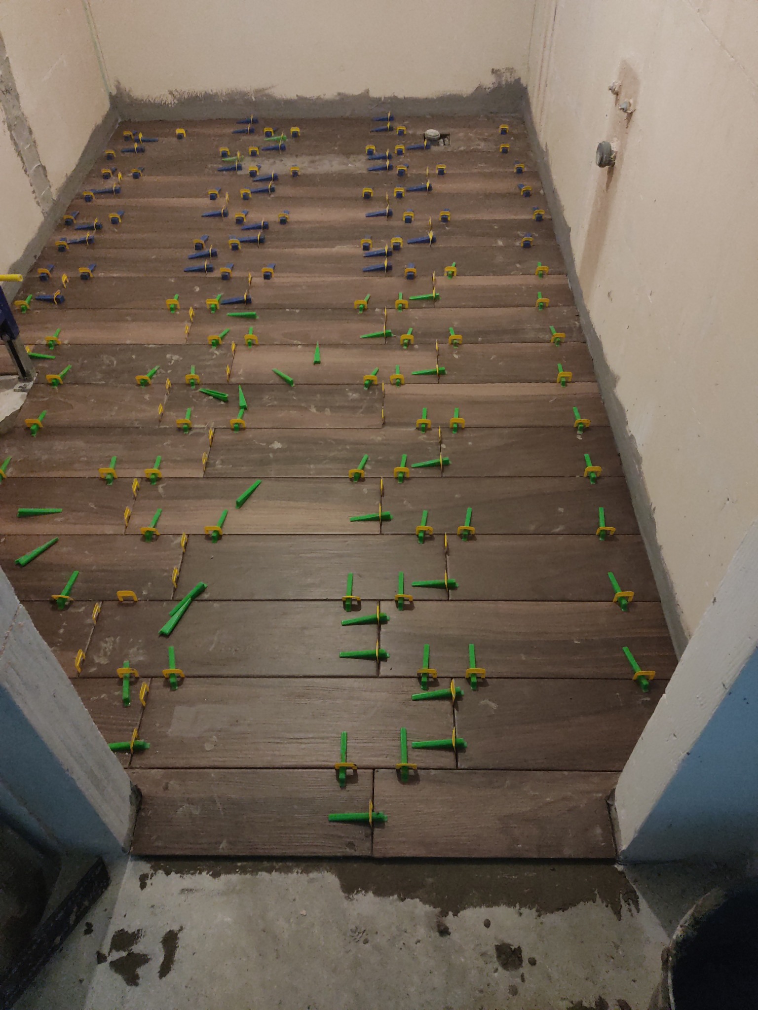 Układanie podłogi z płytek imitujących drewno w Warszawie, z użyciem systemu poziomowania płytek (klipsy i kliny) w trakcie pracy. Widoczne fugi i surowy stan ścian.