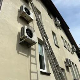 Montaż klimatyzacji na elewacji budynku. Widoczne jednostki zewnętrzne i drabina oparta o ścianę. Ujęcie z dołu podkreśla wysokość instalacji.