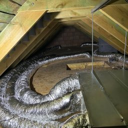MGP Klimatyzacje - Instalacja wentylacyjna na poddaszu: srebrne, elastyczne przewody ułożone między drewnianymi belkami stropu. Widoczny fragment centrali wentylacyjnej i konstrukcja dachu.