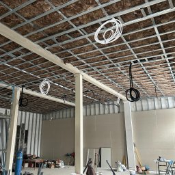 MGP Klimatyzacje - Montaż instalacji pod sufitem podwieszanym z izolacją. Widoczne kable i rury. W tle surowy stan budynku. Wnętrze w trakcie prac instalacyjnych.