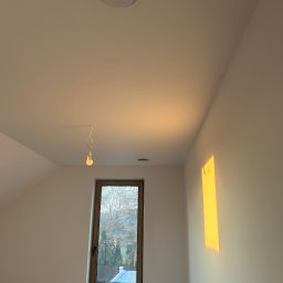 MGP Klimatyzacje - Wnętrze poddasza z oknem, grzejnikiem i okrągłym nawiewnikiem sufitowym. Na suficie widoczna wisząca żarówka. Świecące słońce na ścianie.