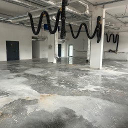 MGP Klimatyzacje - Pusta hala z rozbudowaną instalacją wentylacyjną: srebrne kanały i czarne, faliste przewody pod sufitem. Betonowa podłoga z widocznymi zaciekami.