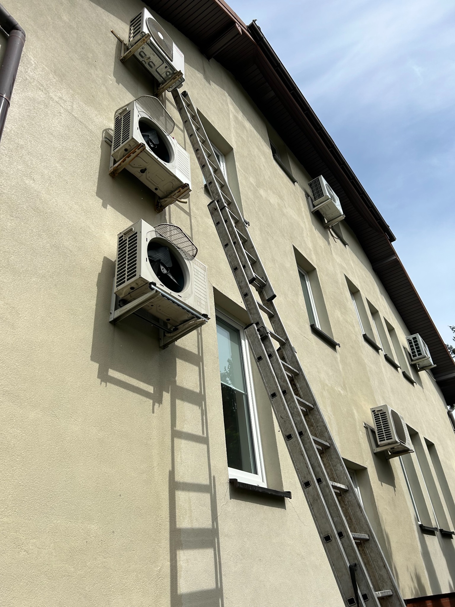 Montaż klimatyzacji na elewacji budynku. Widoczne jednostki zewnętrzne i drabina oparta o ścianę. Ujęcie z dołu podkreśla wysokość instalacji.