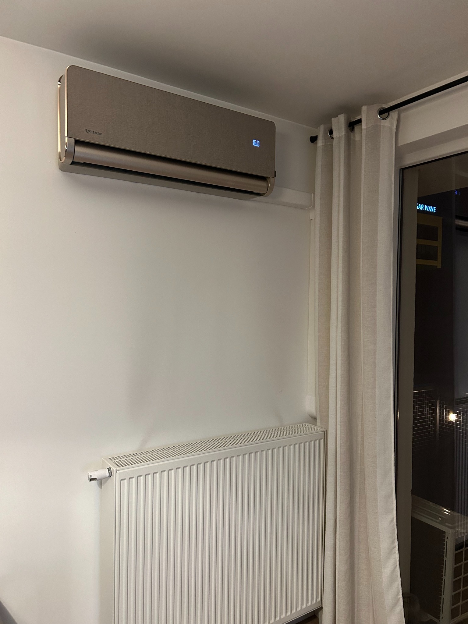 Beżowy klimatyzator Rotenso na ścianie w jasnym wnętrzu. Pod spodem biały grzejnik, obok zasłona przy oknie. Widoczny wyświetlacz temperatury na klimatyzatorze.