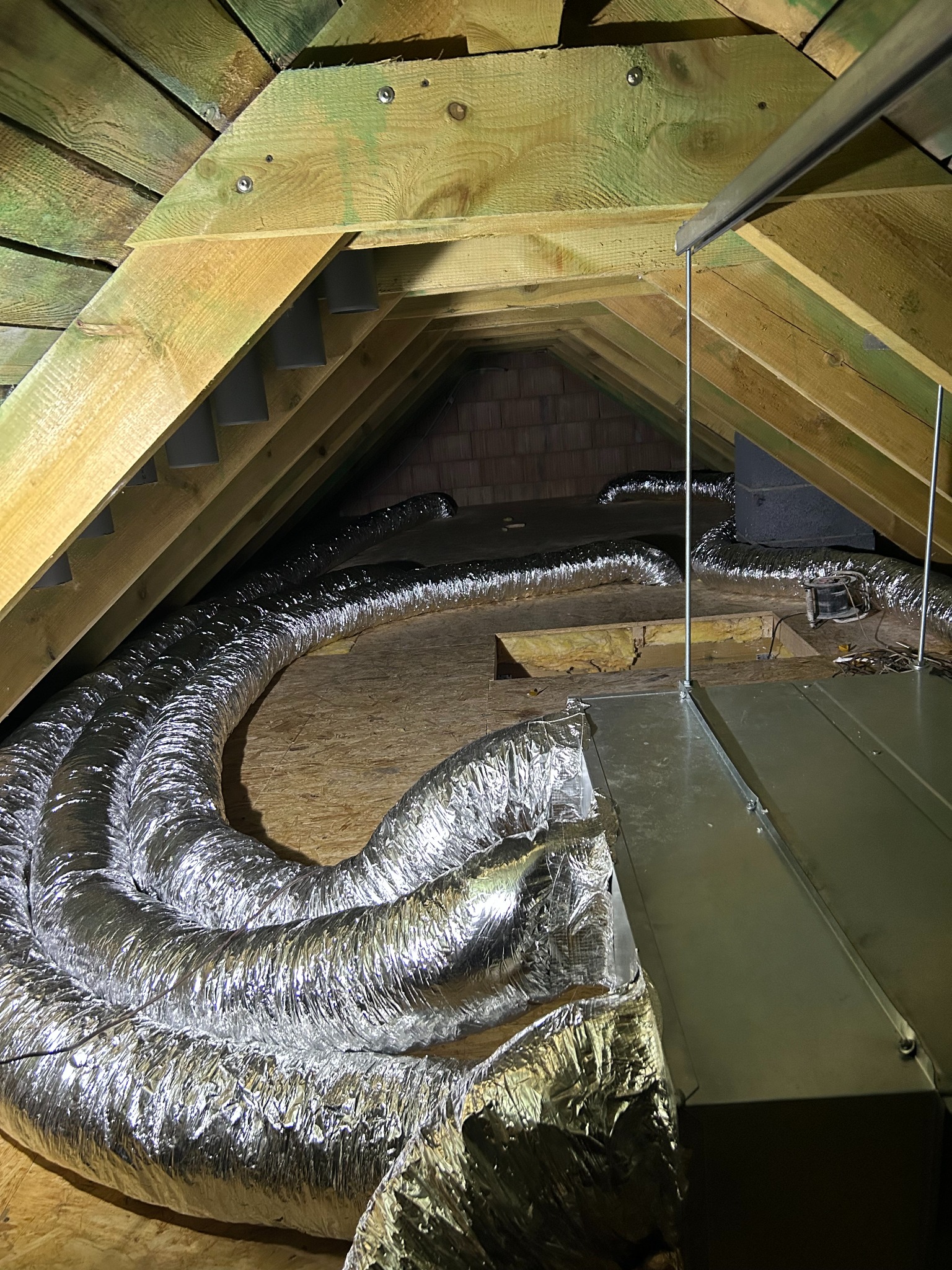 Instalacja wentylacyjna na poddaszu: srebrne, elastyczne przewody ułożone między drewnianymi belkami stropu. Widoczny fragment centrali wentylacyjnej i konstrukcja dachu.