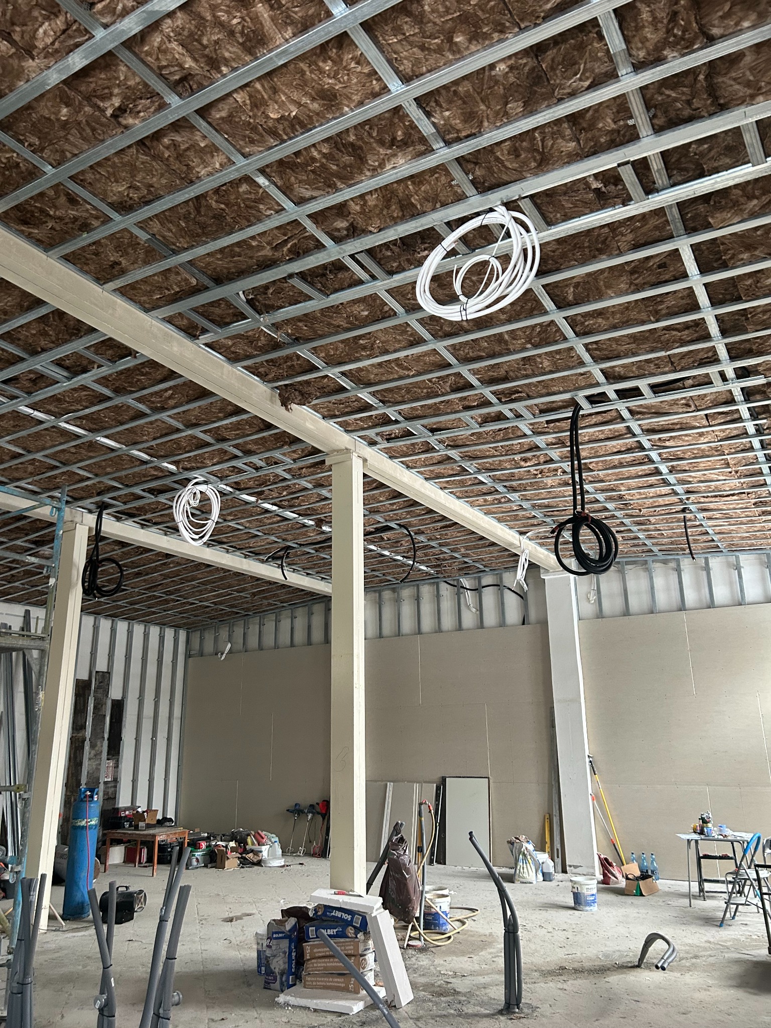 Montaż instalacji pod sufitem podwieszanym z izolacją. Widoczne kable i rury. W tle surowy stan budynku. Wnętrze w trakcie prac instalacyjnych.