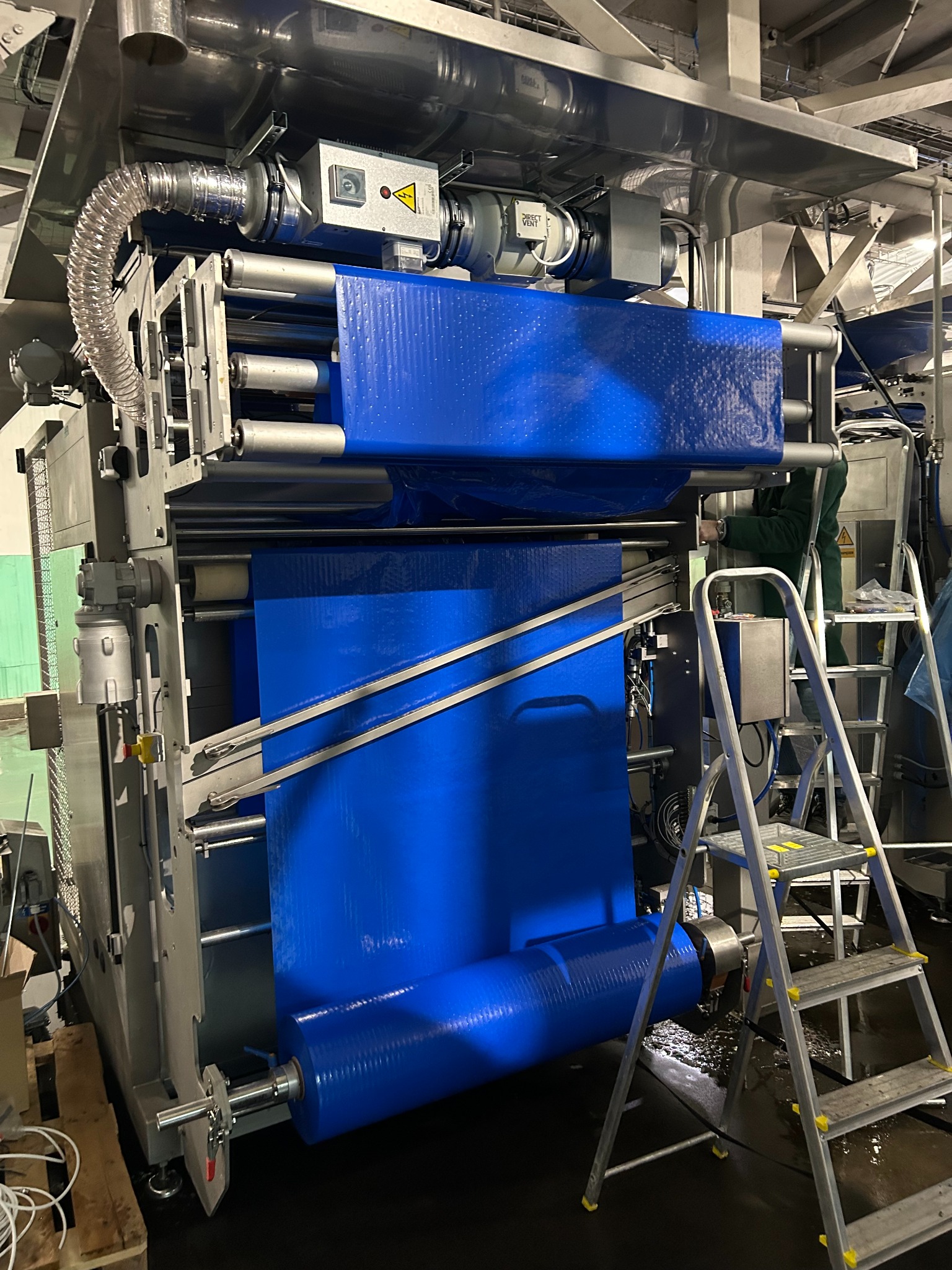 Montaż układu wentylacji przemysłowej z niebieskim filtrem w hali produkcyjnej w Warszawie. Widoczna drabina i pracownik w trakcie instalacji.