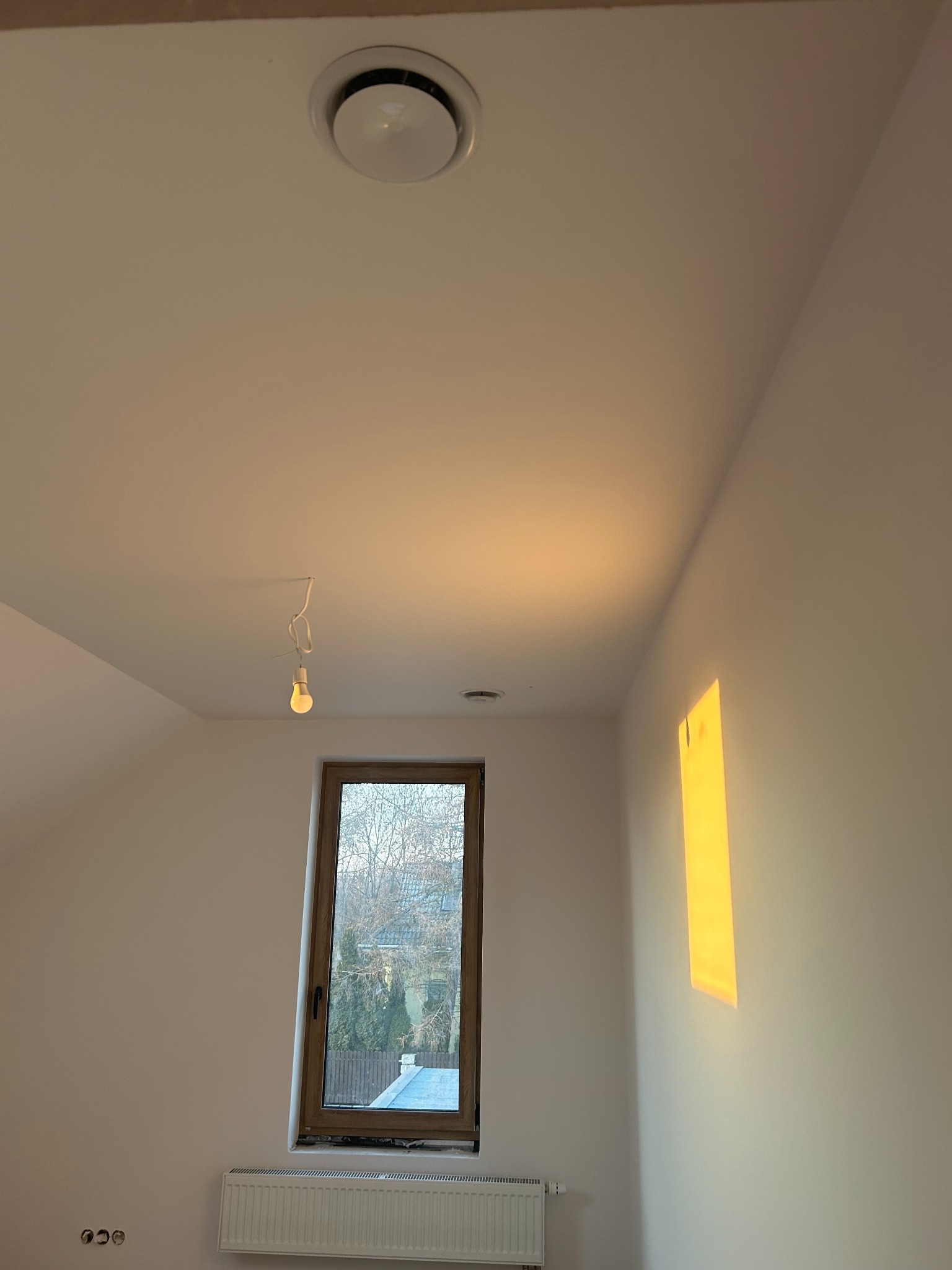 Wnętrze poddasza z oknem, grzejnikiem i okrągłym nawiewnikiem sufitowym. Na suficie widoczna wisząca żarówka. Świecące słońce na ścianie.