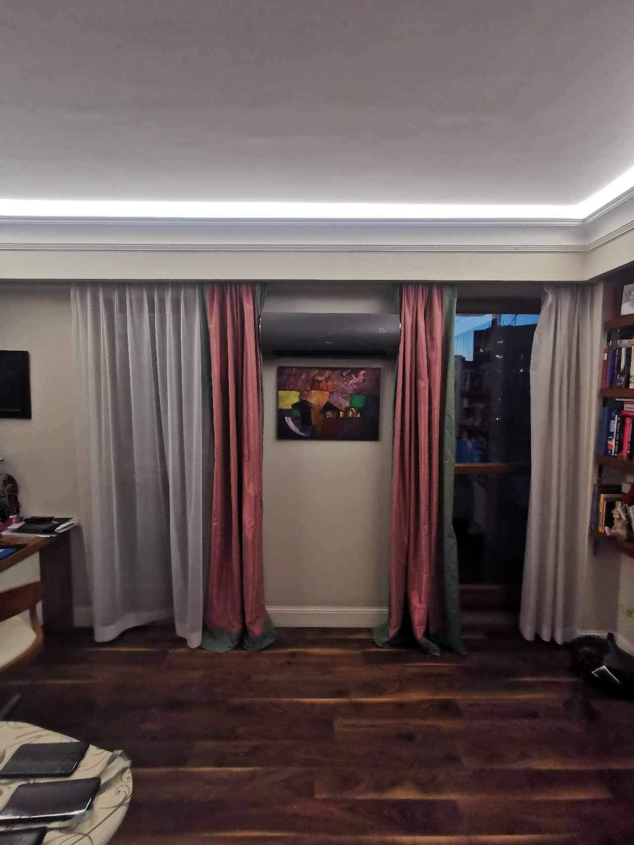 Klimatyzator dyskretnie zamontowany nad obrazem w salonie, zasłonięty częściowo przez różowe zasłony. Widok na budynki za oknem, drewniana podłoga.