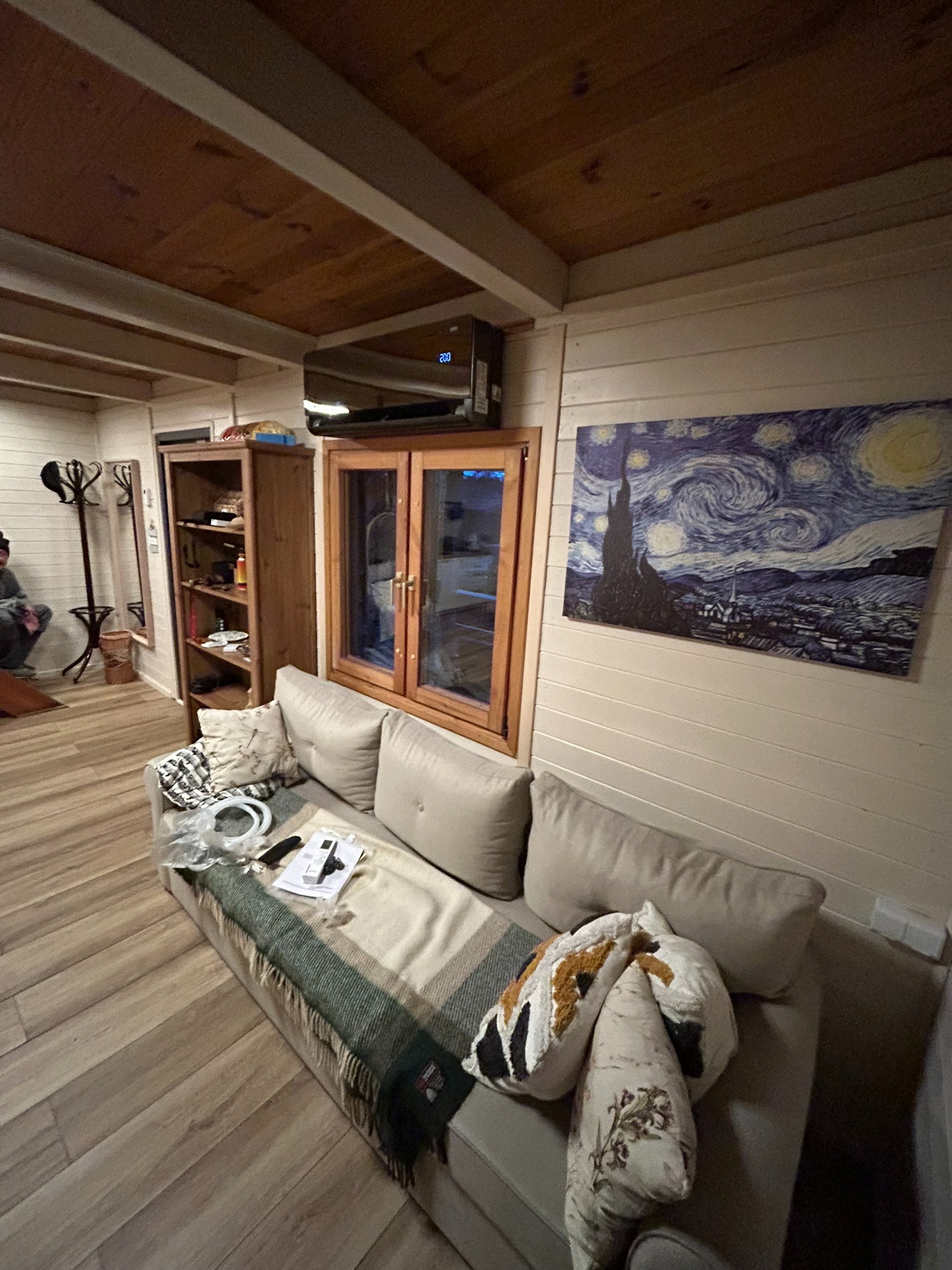Klimatyzator zamontowany nad oknem w drewnianym domku. Na ścianie wisi obraz 'Gwiaździsta noc' van Gogha. Na kanapie leżą piloty i akcesoria.