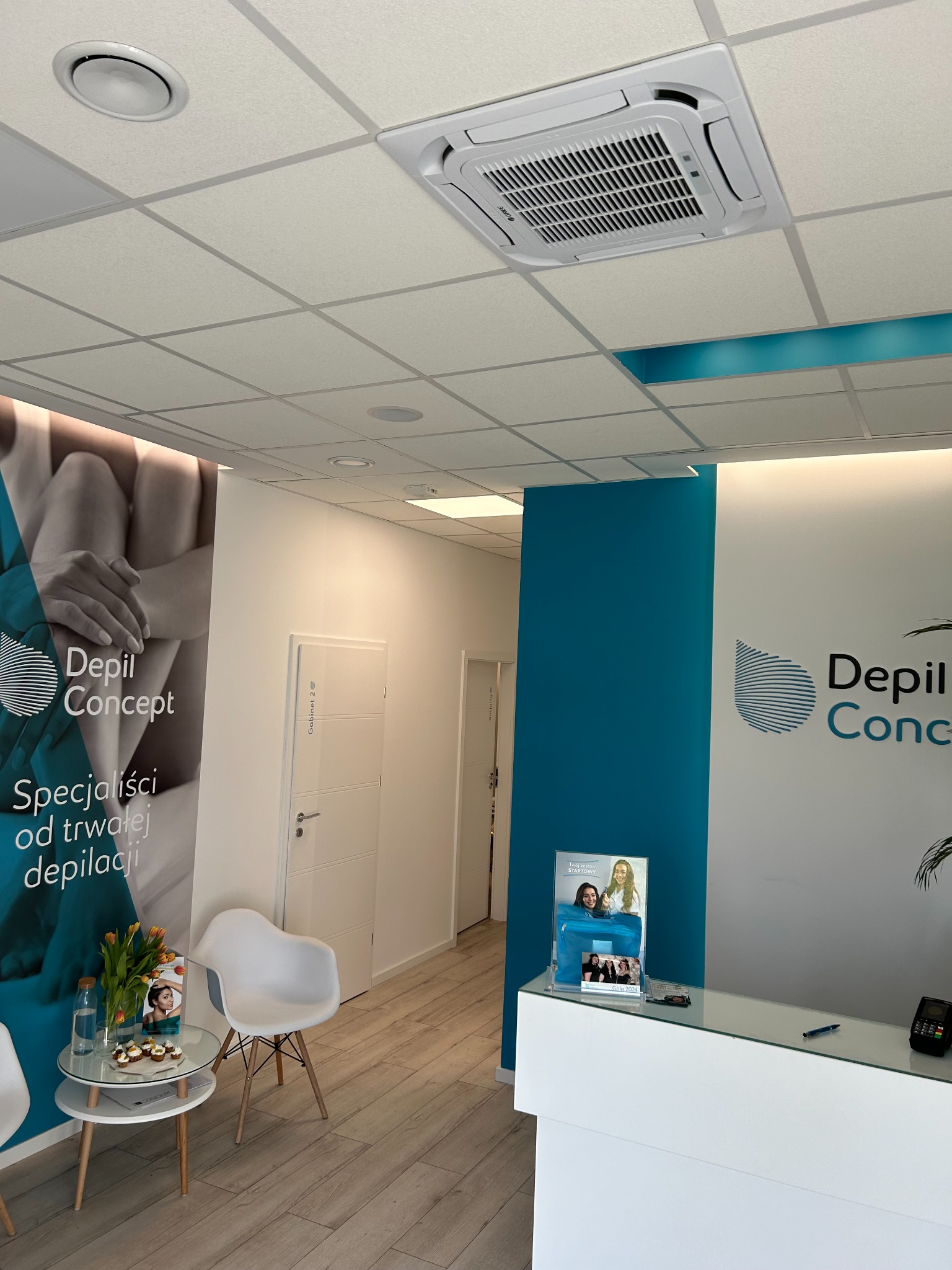 Klimatyzator kasetonowy w nowoczesnym wnętrzu gabinetu kosmetycznego z recepcją i elementami dekoracyjnymi w kolorach bieli i turkusu. Widok na sufit podwieszany.