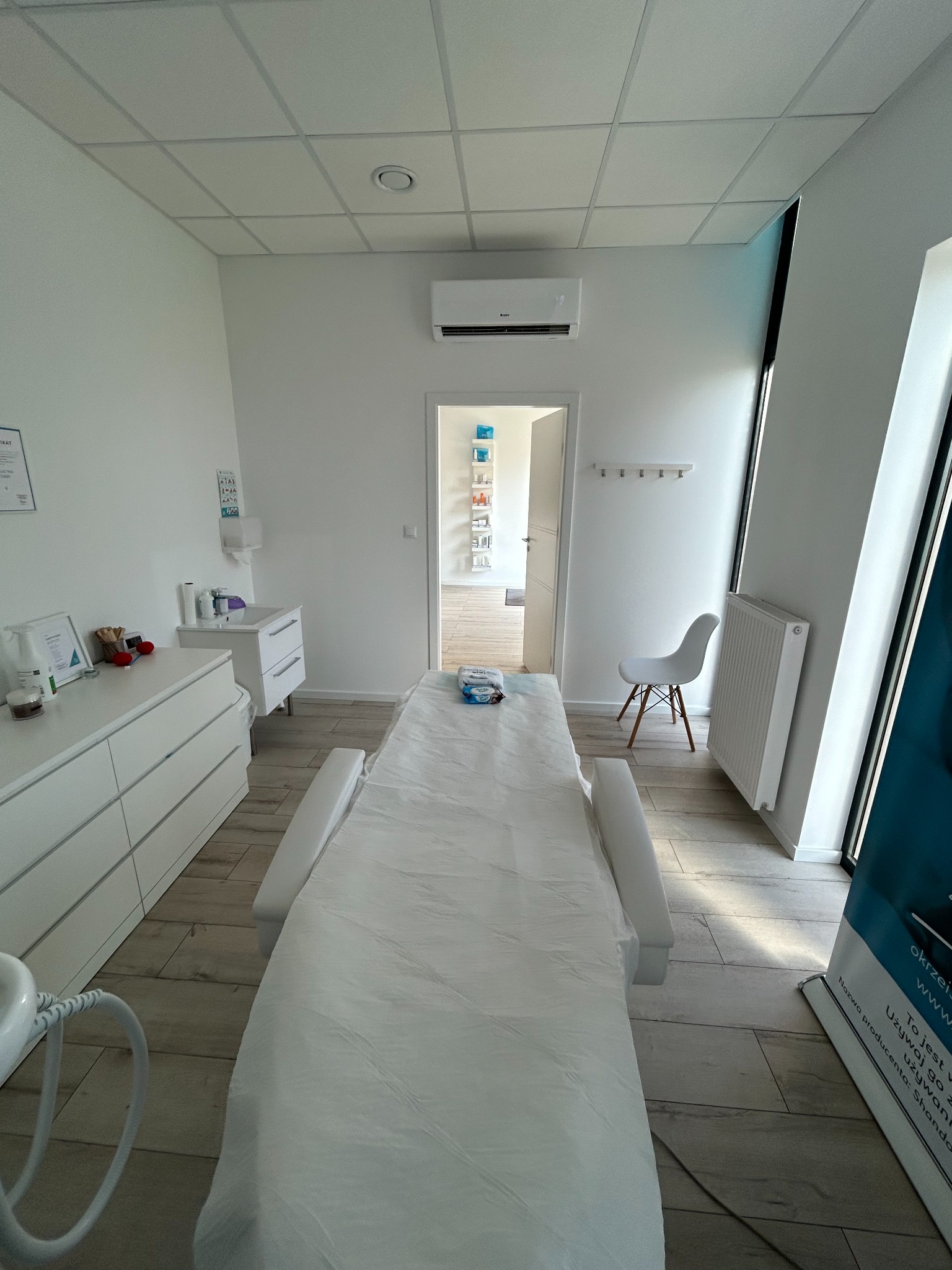 Klimatyzator ścienny w jasnym wnętrzu gabinetu kosmetycznego z leżanką, szafkami i krzesłem. Widoczny również grzejnik i plakat reklamowy.