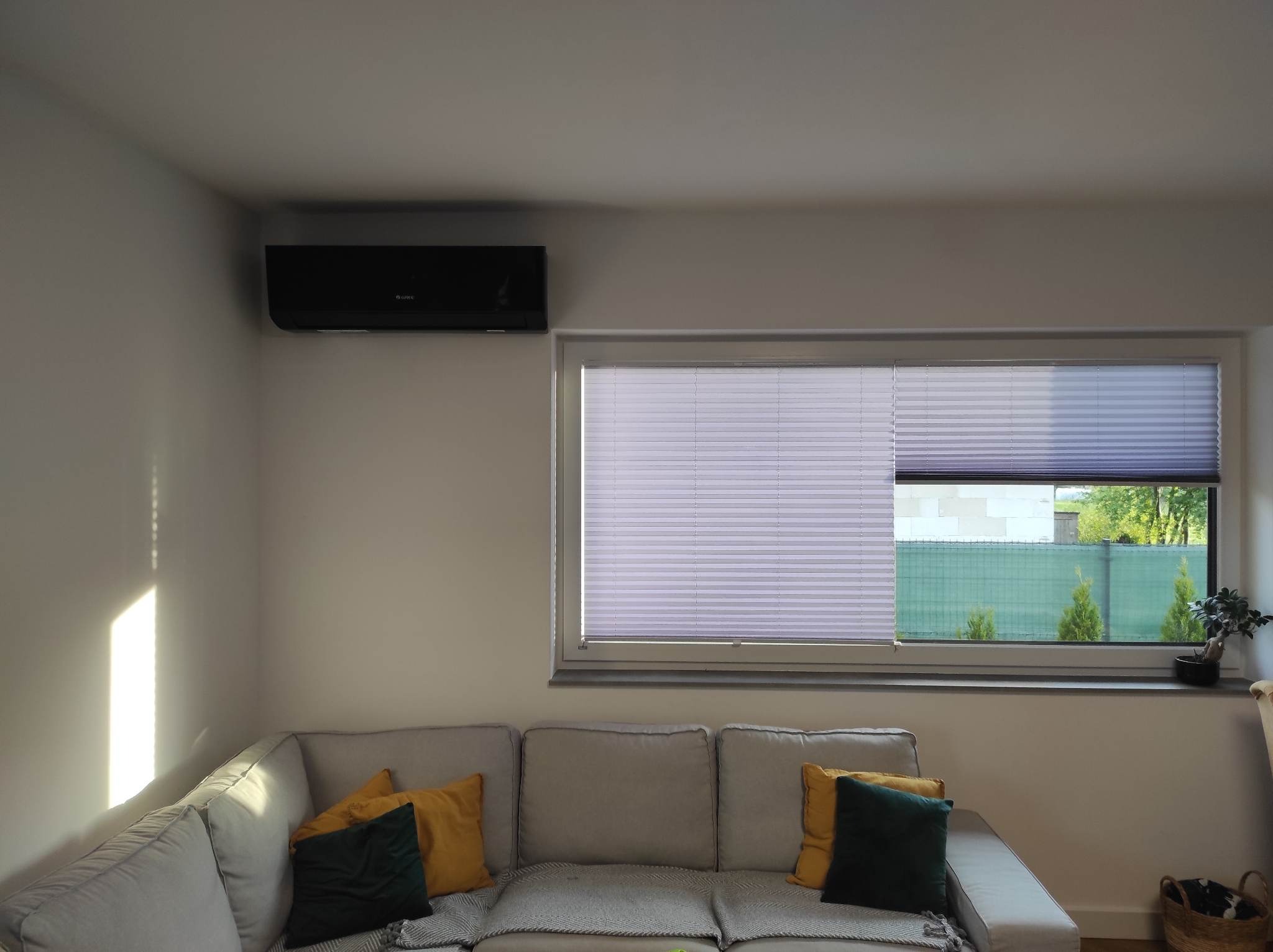 Czarny klimatyzator ścienny w nowoczesnym salonie z szarą sofą, żółtymi i zielonymi poduszkami oraz plisowanymi roletami w oknie.