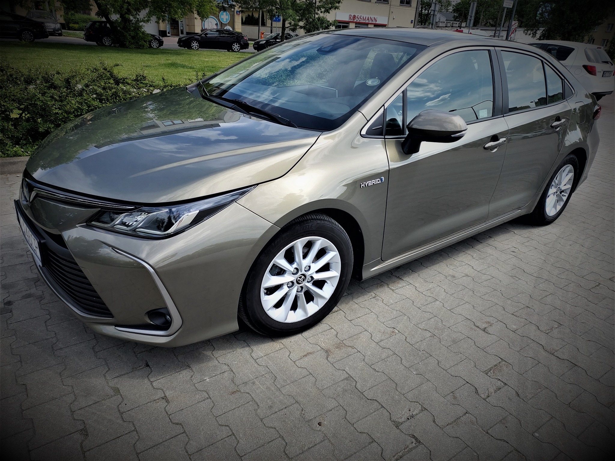 Szaro-złoty samochód osobowy Toyota Corolla Hybrid zaparkowany na chodniku z kostki brukowej, widok z przodu i z boku.