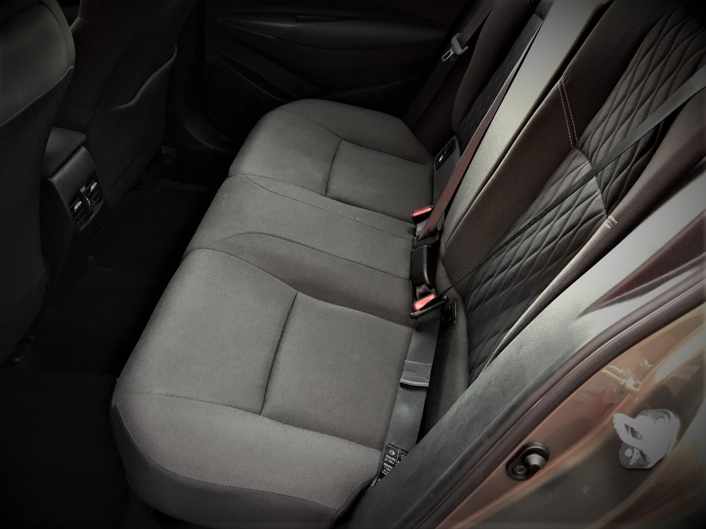 Wnętrze samochodu: tylna kanapa z trzema miejscami, szara tapicerka, pasy bezpieczeństwa z czerwonymi elementami, widoczne kratki wentylacyjne.
