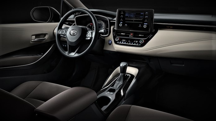 Wnętrze samochodu osobowego z kremową deską rozdzielczą i czarnymi siedzeniami, widoczne kierownica z logo Toyota, ekran dotykowy i automatyczna skrzynia biegów.