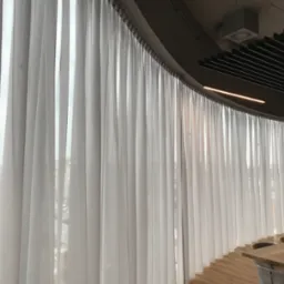 Wysokie, zwiewne, białe zasłony panelowe zakrywające duże okno w nowoczesnym biurze z widoczną strukturą sufitu i elementami oświetlenia liniowego.