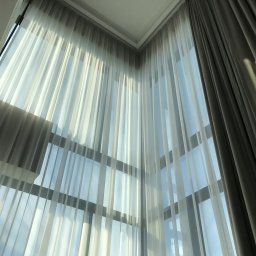 Automatyka zasłon ELEKTRYCZNE.EU - Wysokie okno z delikatnymi, białymi, prześwitującymi firanami i zasłoną w kolorze gołębim, przez które wpada jasne, naturalne światło.