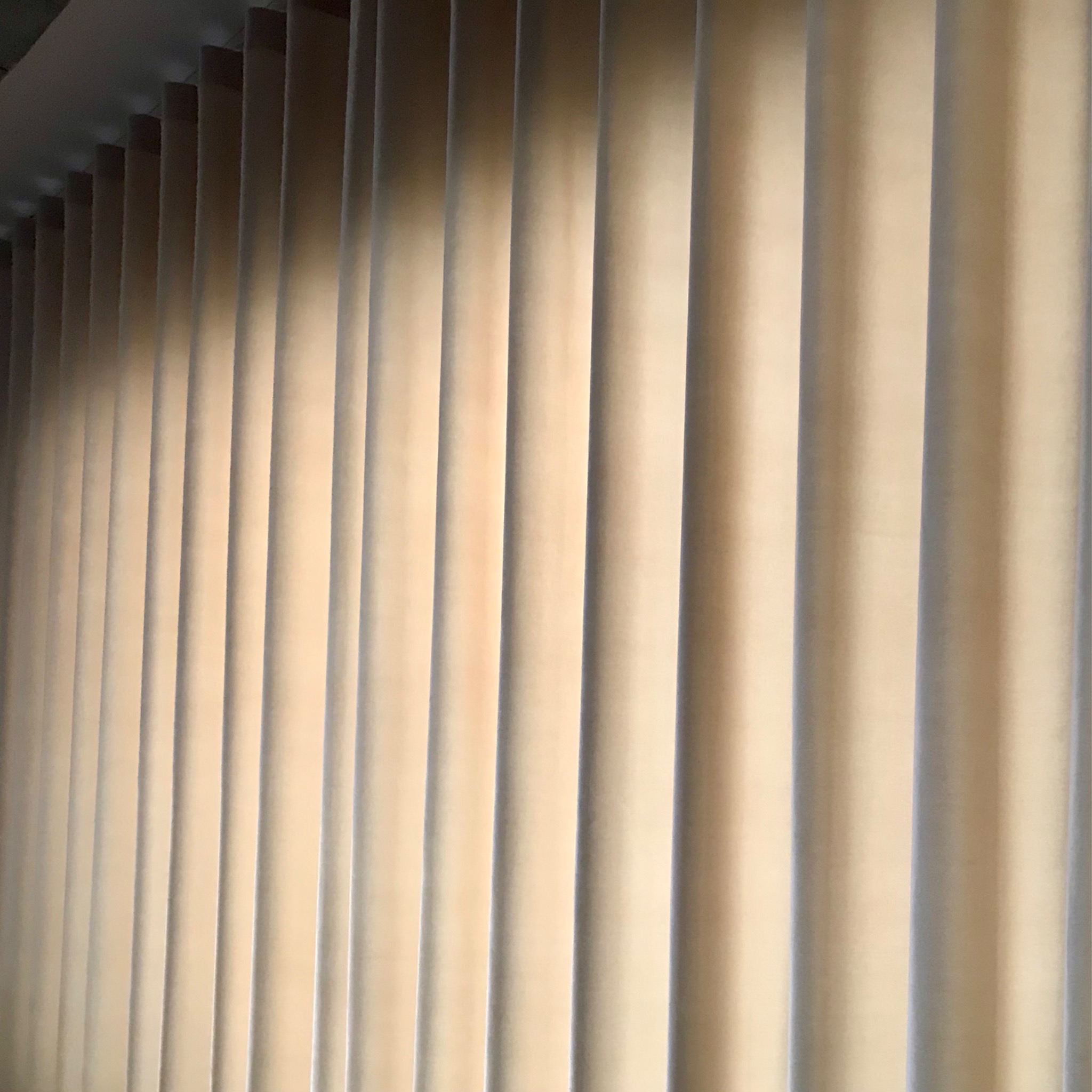 Beżowe, pionowe żaluzje materiałowe, delikatnie pofałdowane, z widocznym cieniem na górze, tworzące rytmiczny wzór.