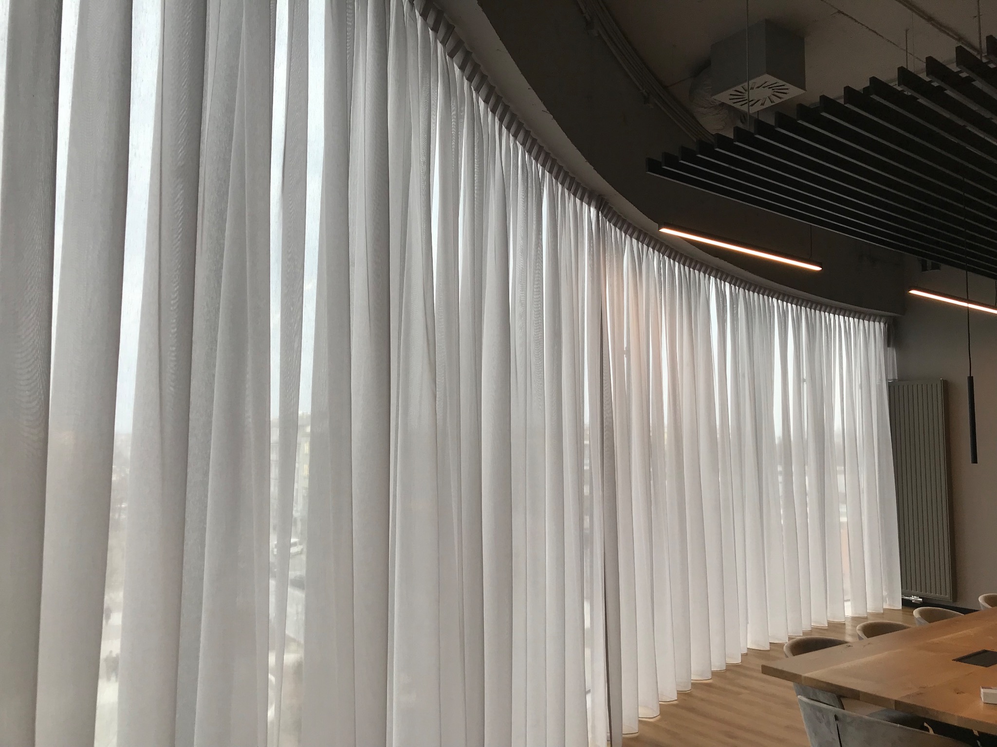 Wysokie, zwiewne, białe zasłony panelowe zakrywające duże okno w nowoczesnym biurze z widoczną strukturą sufitu i elementami oświetlenia liniowego.