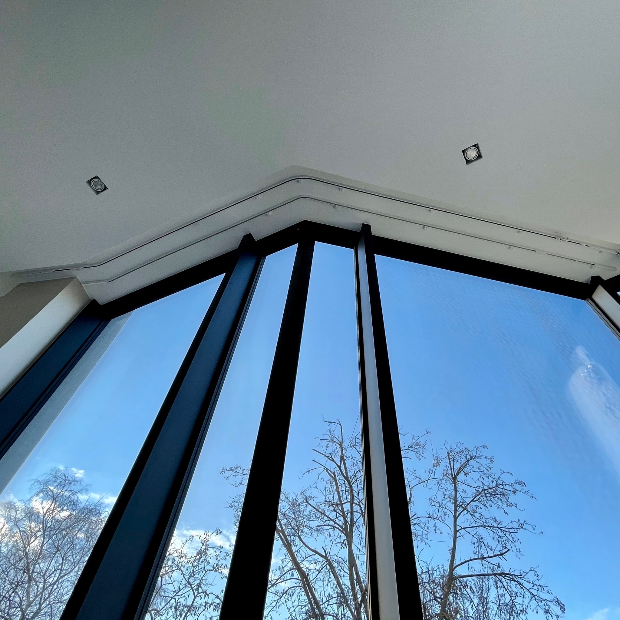 Panoramiczne okno z czarnymi ramami, widok na błękitne niebo i drzewa bez liści, oświetlenie punktowe w suficie