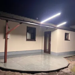 Elewacja domu jednorodzinnego w nocy, oświetlona dwoma lampami LED, z nową, szarą, ceramiczną podłogą tarasu i świeżo pomalowaną fasadą w kolorze beżowym.